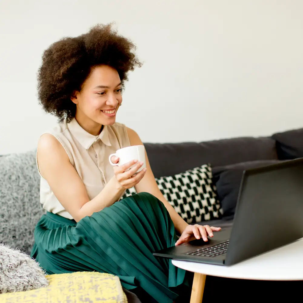 Woman on laptop taking virtual psychotherapy smiling with cup in hand